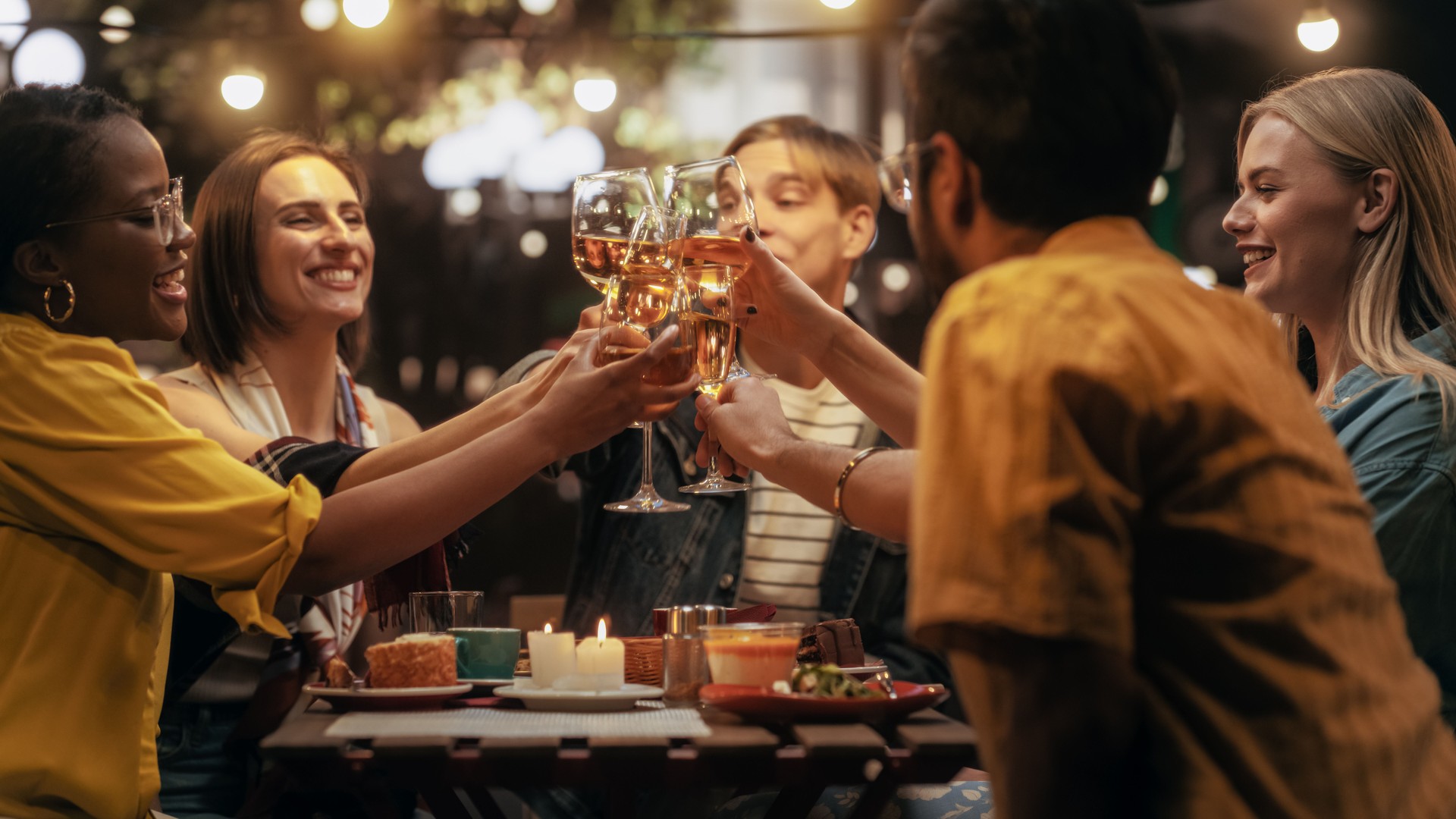 Multiethnic Diverse Friends Gathering Together at a Cafe Terrace in the Evening. Diverse Young People Raising, Toasting and Clinking Glasses with Wine and Champagne, Celebrating Their Reunion